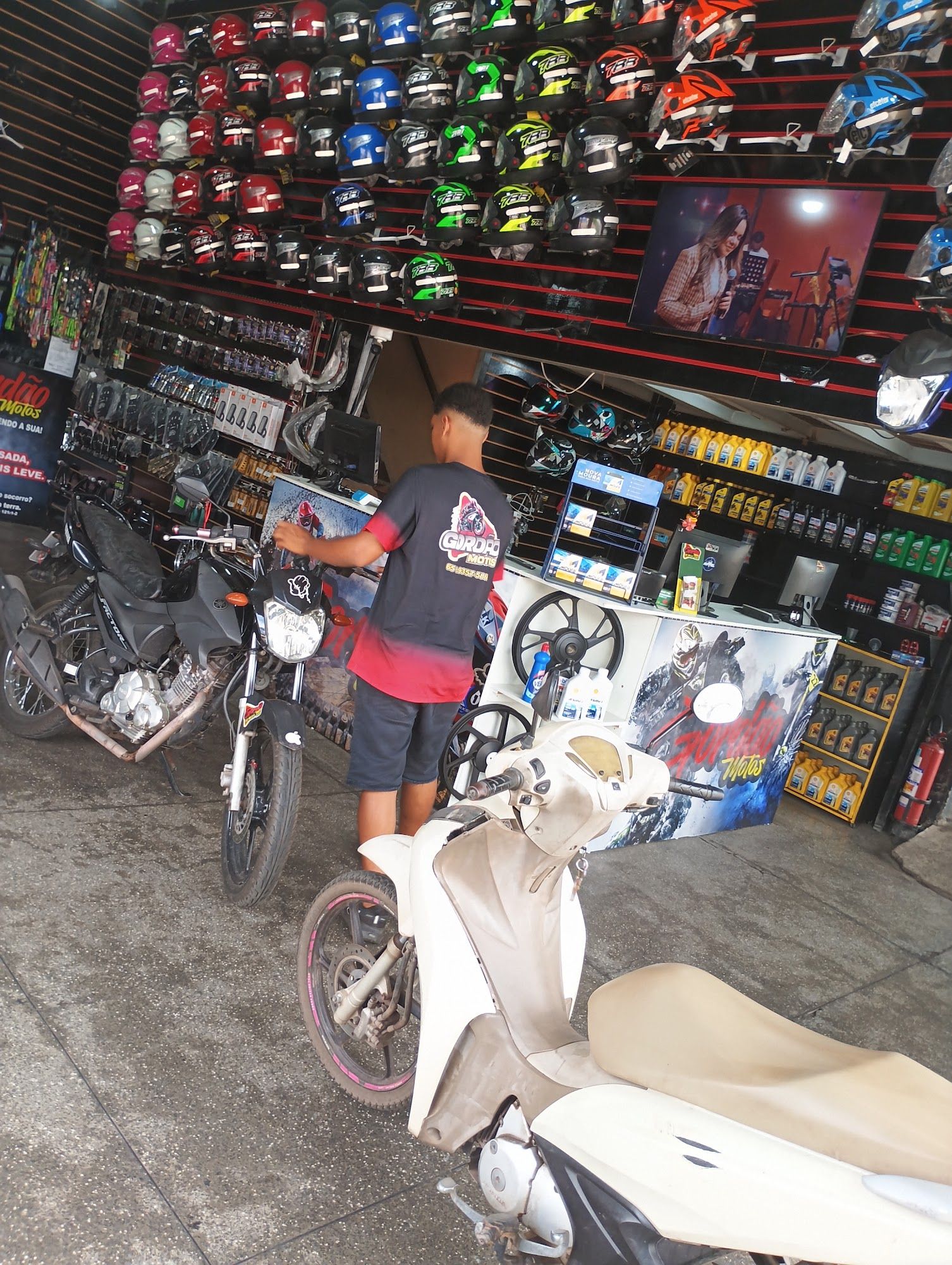 Rapaz com camiseta da Gordão no balcão, com moto e scooter na frente e parede de capacetes ao fundo